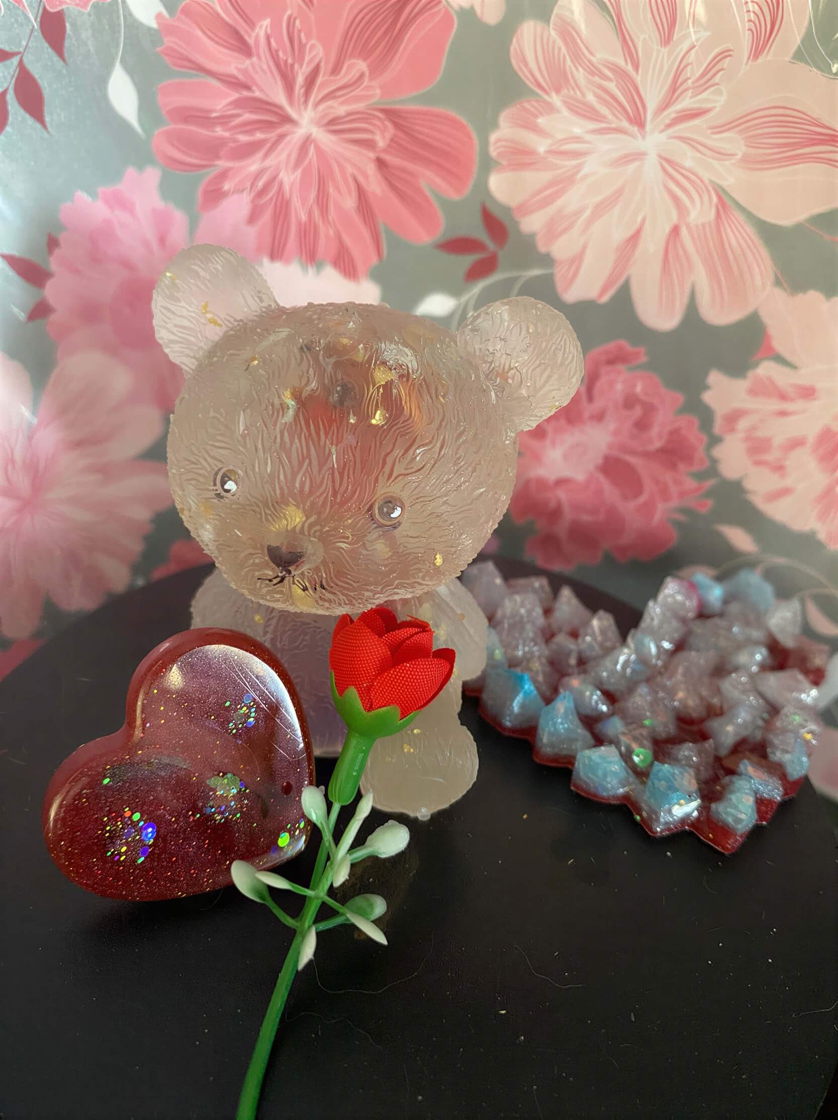 Clear bear-shaped figurine with a rose and heart-shaped glittery decorations on a black surface, floral background.