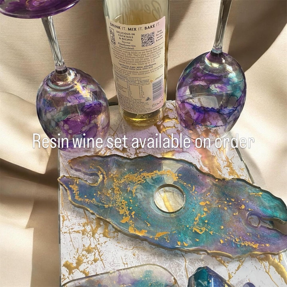 Two purple and teal resin wine glasses and a matching tray with gold flakes next to a glass bottle.