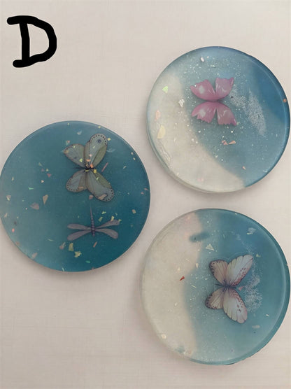 Set of three round coasters featuring embedded butterfly designs in teal and clear resin with glitter accents.