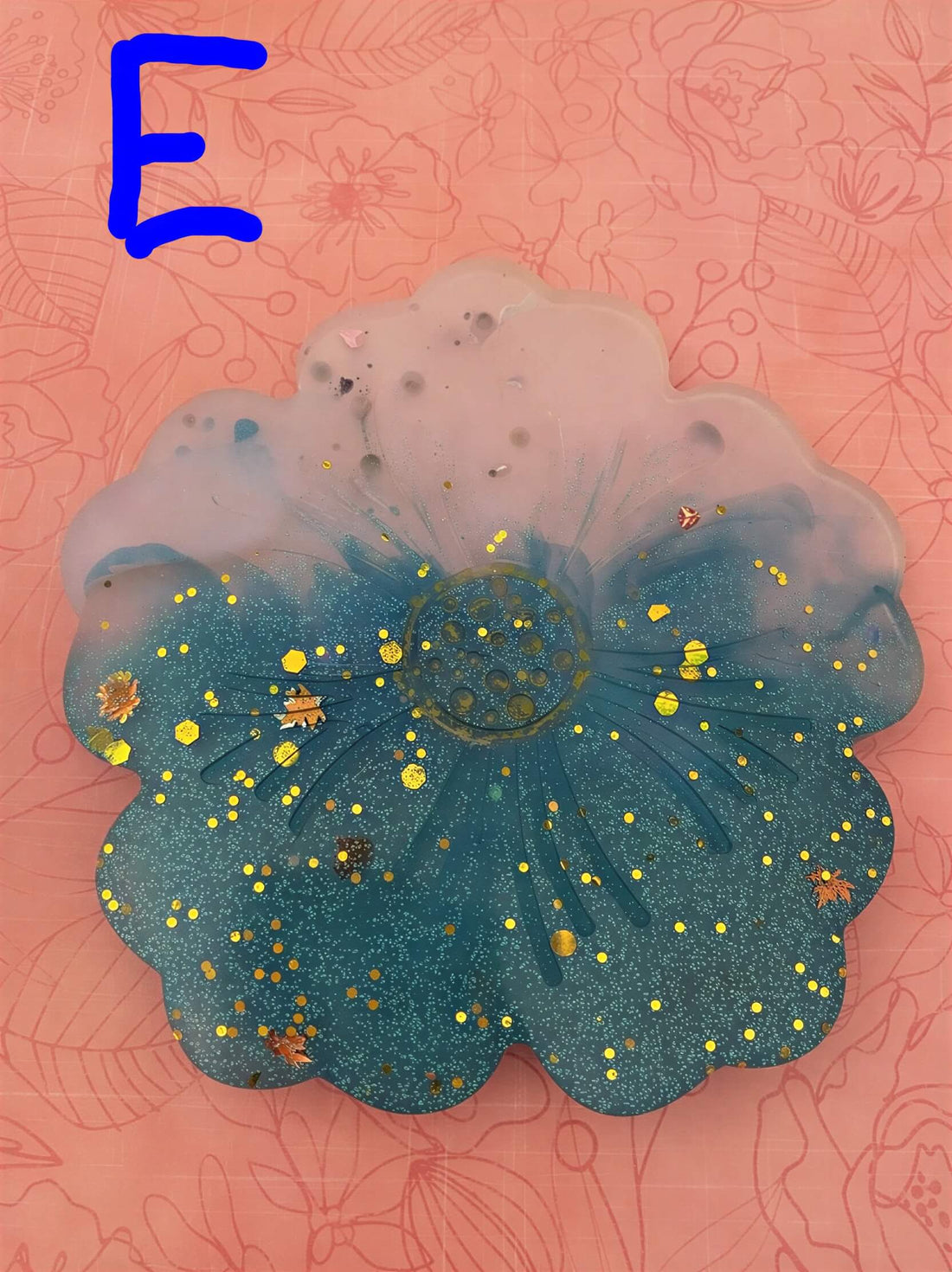 Flower-shaped coaster in teal with gold specks, set against a pink floral background.