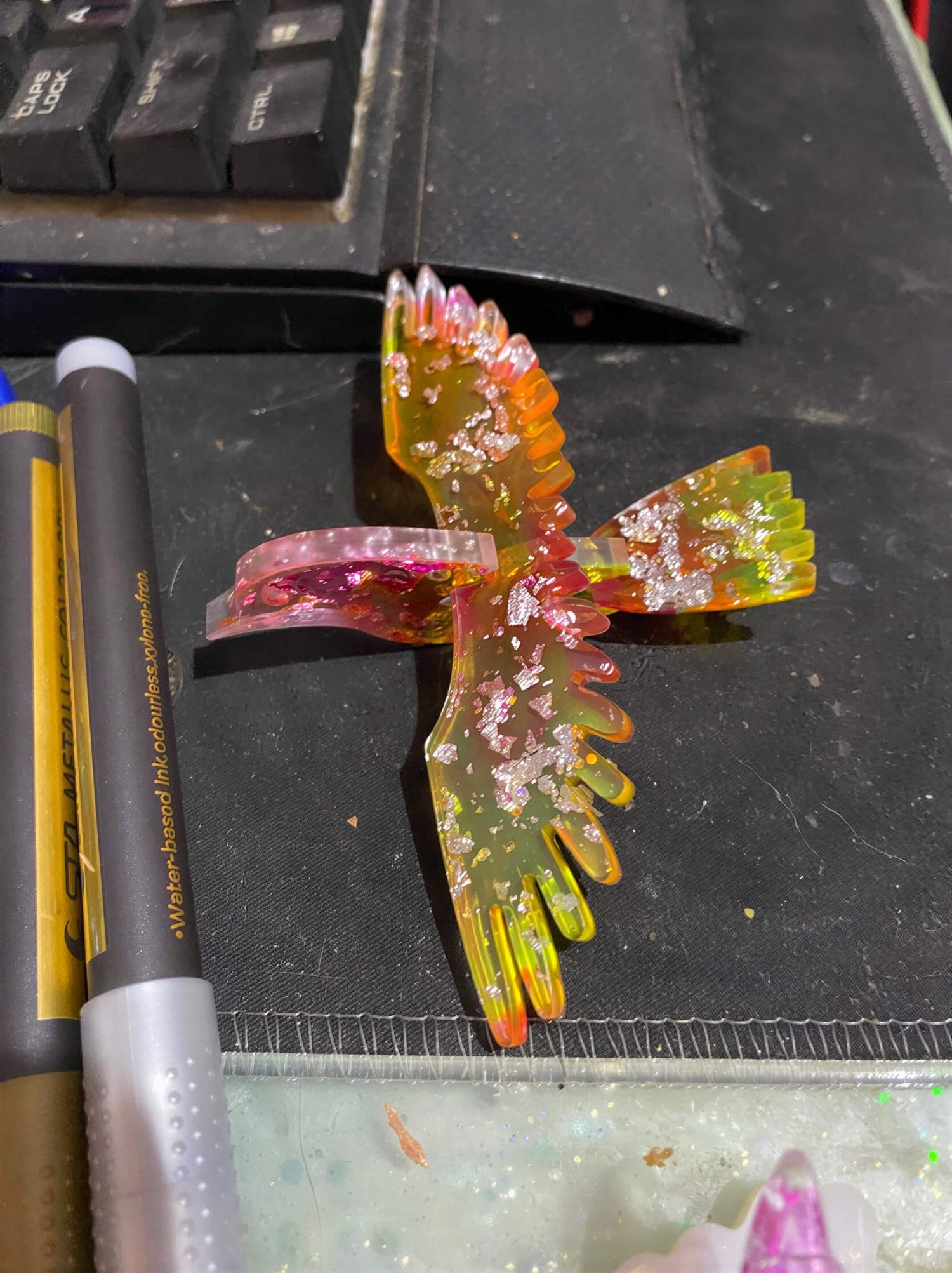 Colorful resin bird sculpture with glitter, positioned on a black surface near pens and a keyboard.