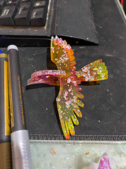 Colorful resin bird sculpture with glitter, positioned on a black surface near pens and a keyboard.