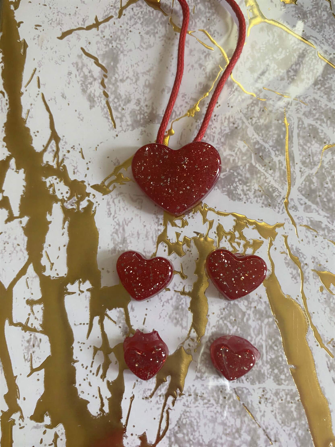 Red heart-shaped jewelry set with a larger pendant and four smaller charms, featuring a glittery finish on a gold marbled ...