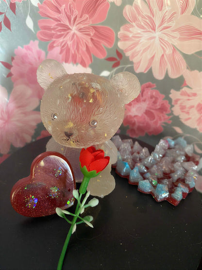 Clear bear-shaped figurine with a rose and heart-shaped glittery decorations on a black surface, floral background.