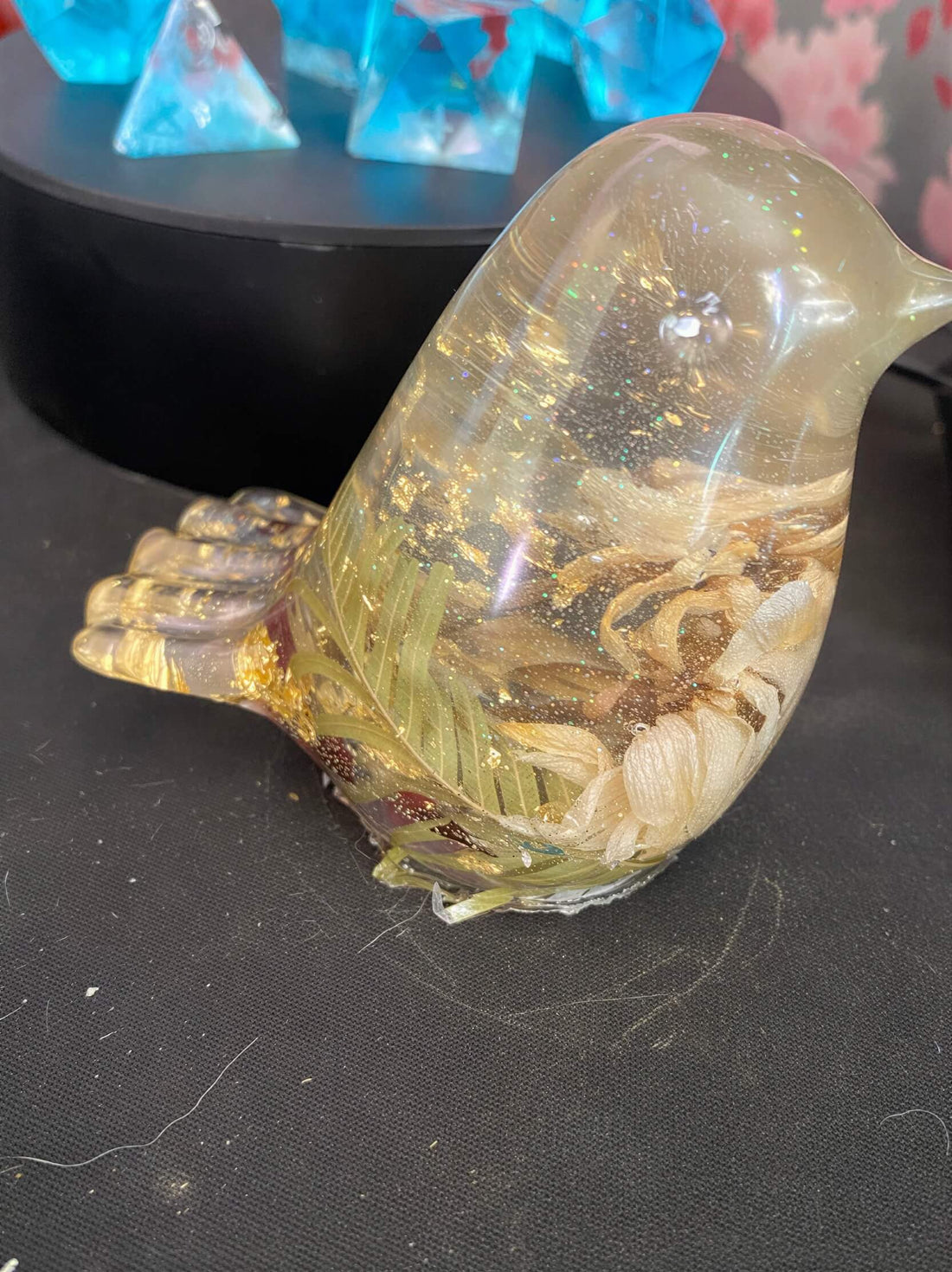 Clear resin bird sculpture with gold flakes and floral elements, positioned on a dark surface.