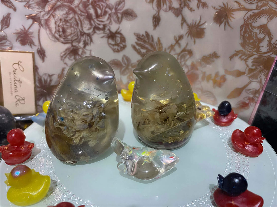 Worry Bird Pair in resin, featuring two rounded birds with embedded natural elements, surrounded by colorful decorative it...