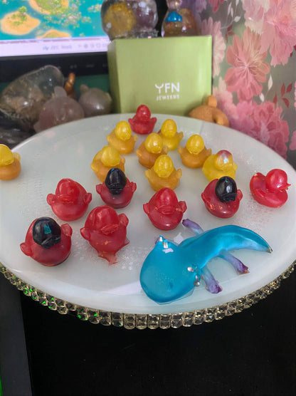 Colorful resin ducks in various poses on a white plate, with a blue whale figurine and decorative stones in the background.